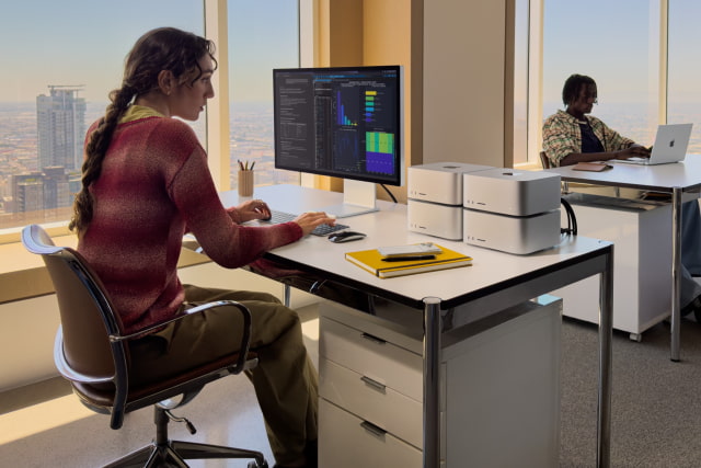 A person in an office sits in front of a desk that has a large display connected to four Mac Studio devices.