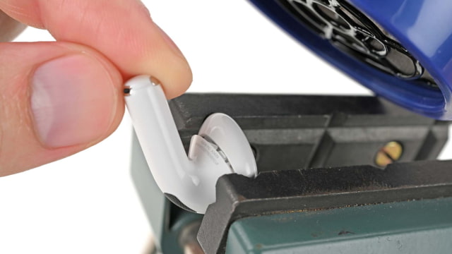 White AirPod secured in a vise under a blue hair dryer with a hand holding the stem